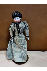 Puppe mit Brustblattkopf aus glasiertem weißen Porzellan mit modellierten schwarzen Haaren und gemalten blauen Augen, Stoffkörper, Arme aus Porzellan. Wohl um 1880, ohne Marke. Auf Metallständer montiert.