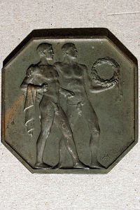 Achteckige Plakette: 2 antike Athleten einander umarmend, einer mit Lorbeerkranz, der andere mit Tuch.